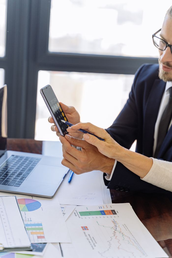 gallery-4 Business professionals reviewing financial reports on a smartphone at a desk with papers and a laptop in an office setting.